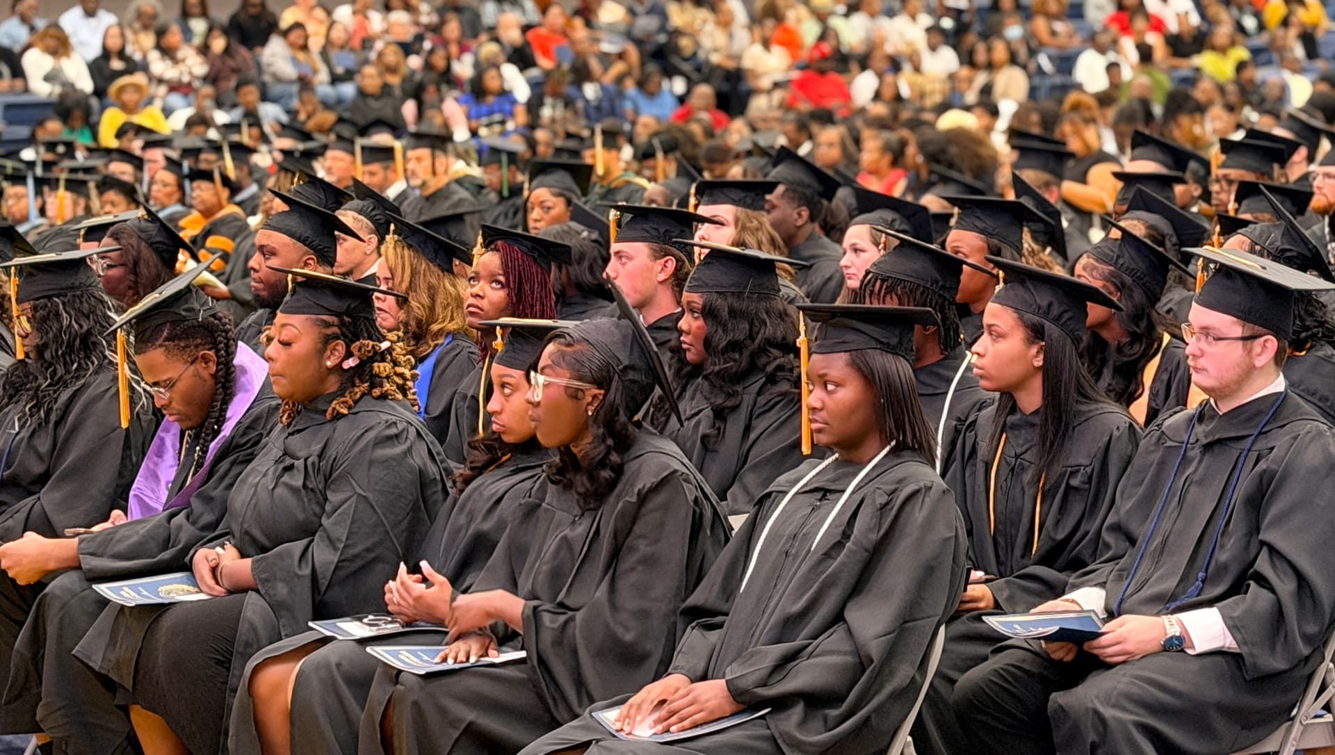 Students in black caps and gowns listen to speakers onstage at graduation